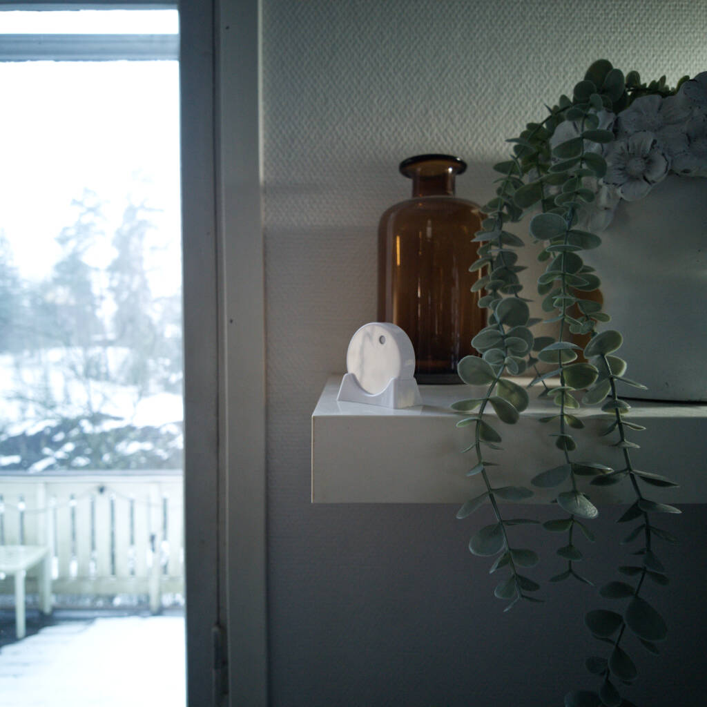 RuuviTag sensor on a living room shelf with a brown glass vase, green plant, and snowy outdoor view.