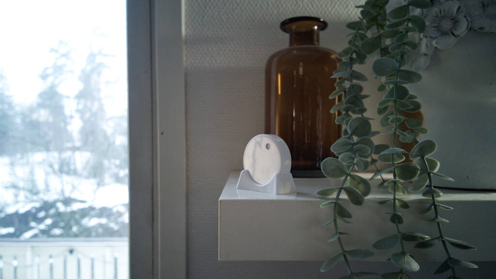 RuuviTag sensor on a living room shelf with a brown glass vase, green plant, and snowy outdoor view.
