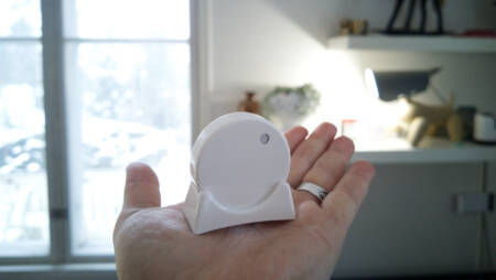A person holding a small, round RuuviTag sensor in their palm, with a bright living room and window in the background.