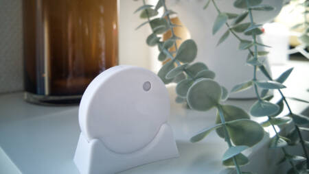 Minimalist white RuuviTag sensor on a shelf, next to a brown glass vase and a leafy plant in a white decorative pot.