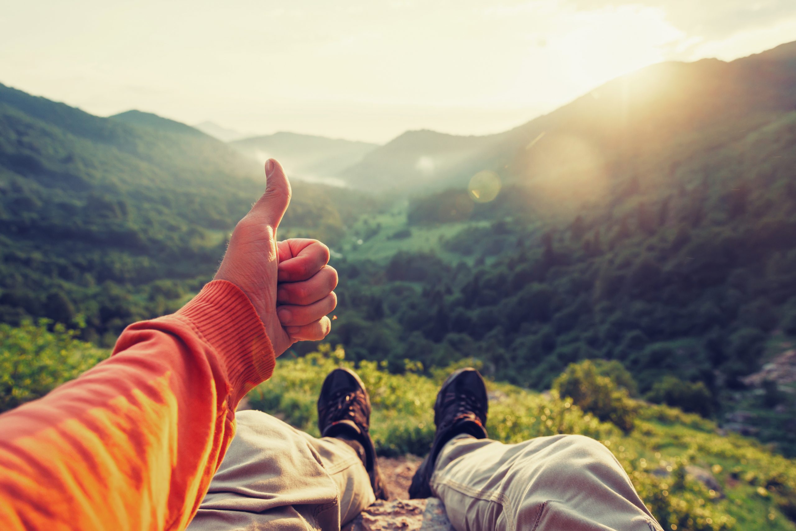 Happy male traveler showing thumb up in summer mountains at sunset, point of view shot