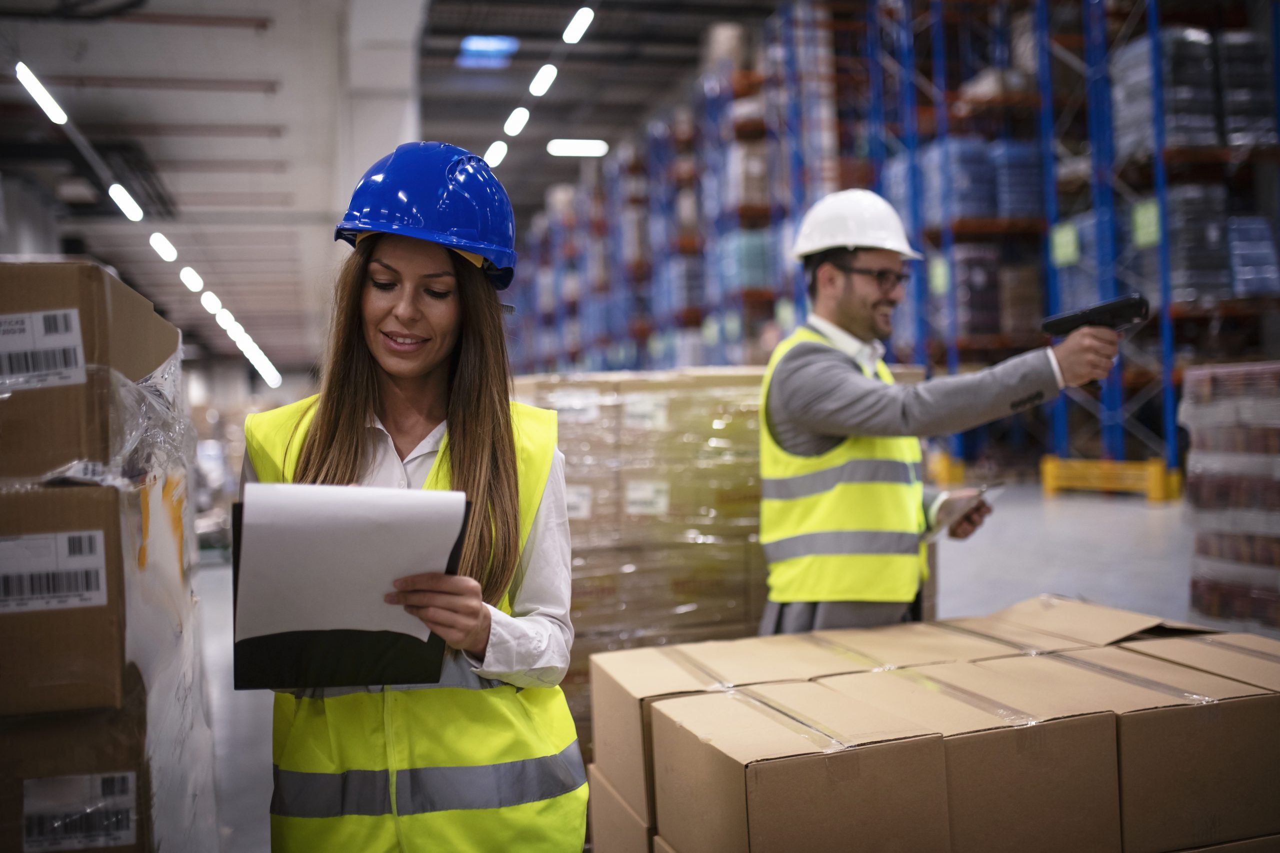 Female factory worker in reflective uniform with hardhat helmet checking new arrival of goods in warehouse while worker using bar code reader in background. Logistics and distribution.