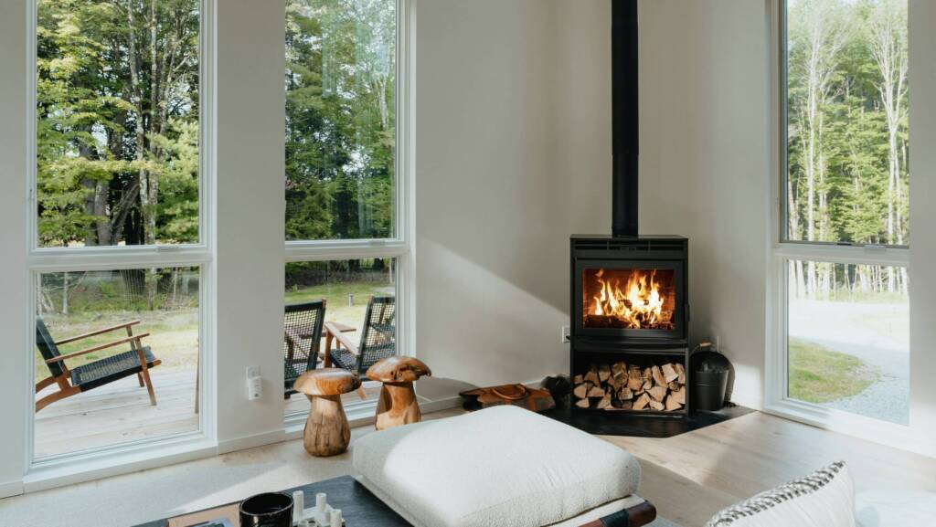 Bright living room with large windows, modern wood stove, stacked firewood, and a view of green trees outside.