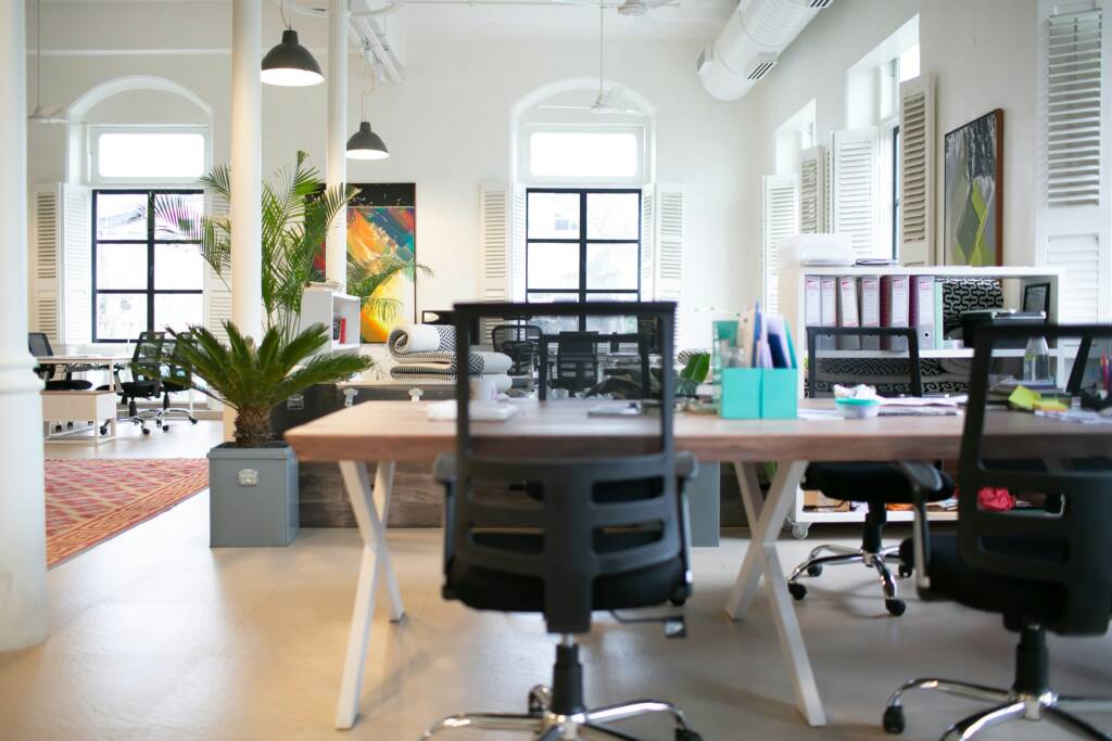 Modern open-plan office with ergonomic chairs, large windows, indoor plants, and bright natural lighting.
