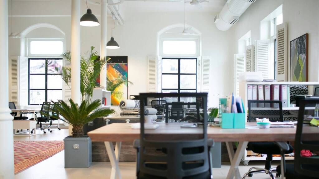 Modern open-plan office with ergonomic chairs, large windows, indoor plants, and bright natural lighting.