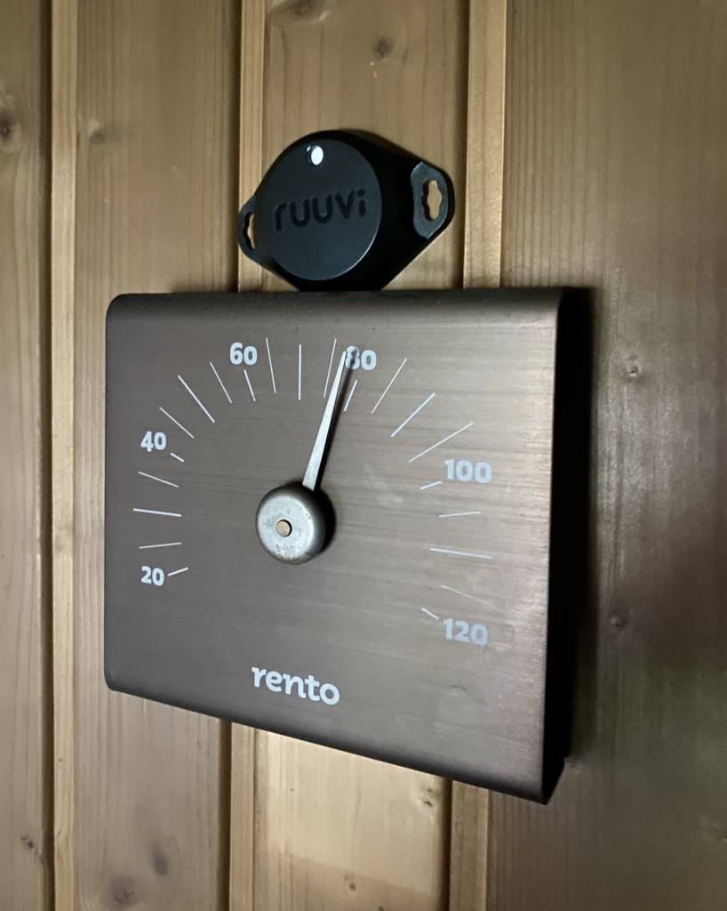 A smart sensor Ruuvitag placed on top of a traditional sauna thermometer in a sauna.