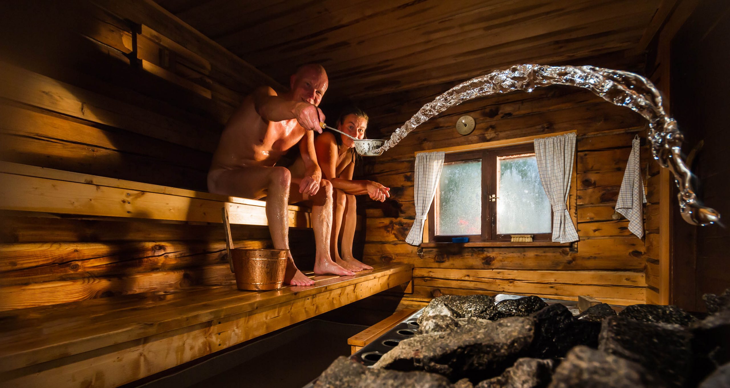 Middle aged couple in traditional wooden Finnish sauna, man throwing water to hot stove